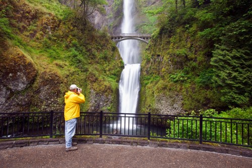 A person in a yellow jacket photographs a waterfall surrounded by lush greenery and a bridge above.