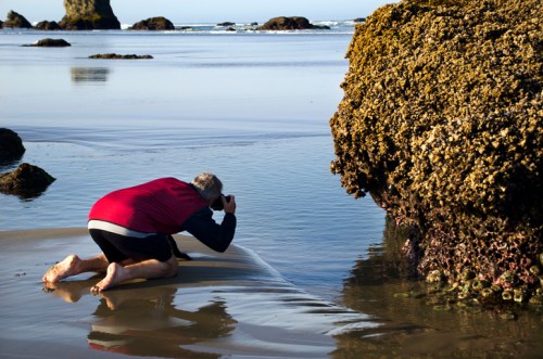 A person kneels on a beach, photographing a rock formation with tide pools, surrounded by calm water and distant waves.