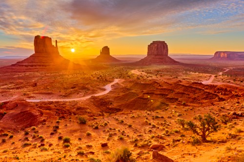 Sunrise over Monument Valley, highlighting iconic rock formations and a winding dirt road in a desert landscape.