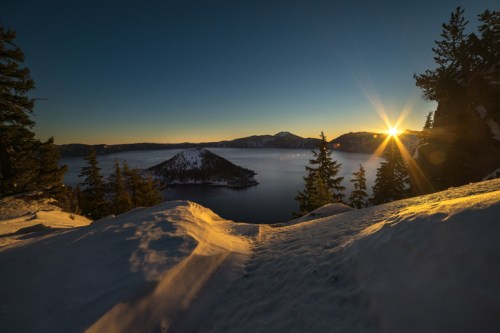 A serene winter landscape at sunrise, featuring a snowy foreground, a lake, and a distant island surrounded by mountains.