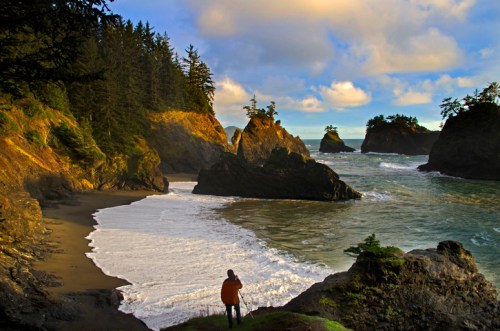 A person stands on a beach, gazing at rocky cliffs and waves under a partly cloudy sky.