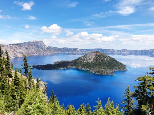 A scenic view of Crater Lake, featuring a blue lake surrounded by trees and a small island under a partly cloudy sky.