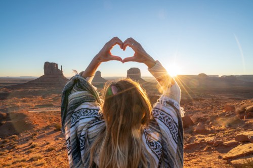 A person in a patterned poncho forms a heart shape with their hands against a sunset over a desert landscape.
