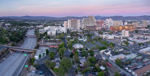 Aerial view of a cityscape at dusk, featuring buildings, highways, and mountains in the background.