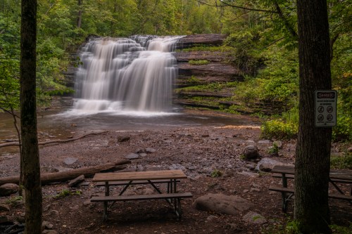 A serene waterfall cascades over rocks, surrounded by lush greenery, with picnic tables in the foreground.