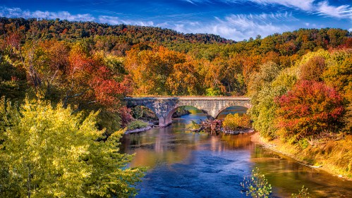 A scenic view of a stone bridge over a calm river, surrounded by vibrant autumn foliage and a clear blue sky.