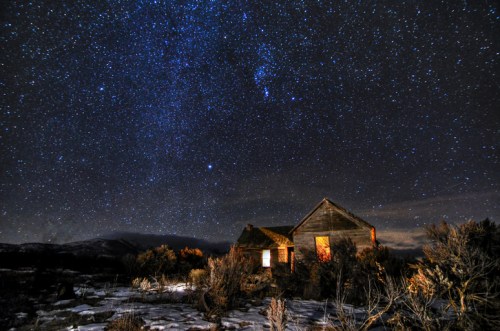 A starry night sky above an old, abandoned house surrounded by sparse vegetation.