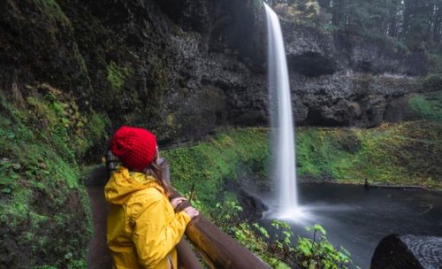 A person in a yellow raincoat and red hat gazes at a tall waterfall surrounded by lush greenery.