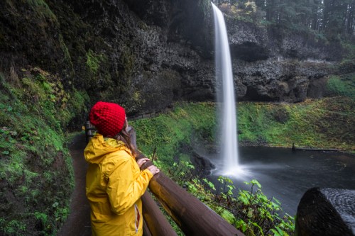 A person in a yellow raincoat and red hat gazes at a tall waterfall surrounded by lush greenery.