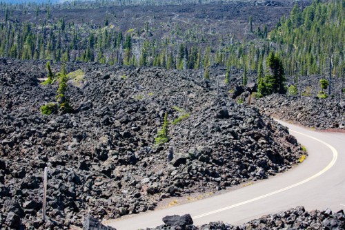 A winding road through a rugged landscape of volcanic rock and scattered pine trees under a clear blue sky.