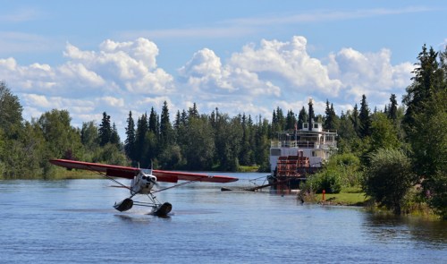 A seaplane takes off over a river, with a vintage boat docked nearby and lush trees lining the shore under a blue sky.