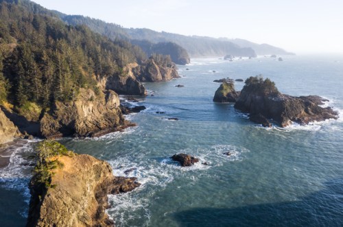 Aerial view of rugged coastline with rocky outcrops, lush forests, and waves crashing against the shore.