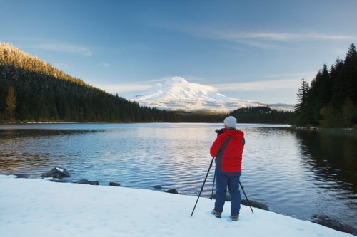 A person in a red jacket stands by a snowy lake, photographing a snow-capped mountain in the background.