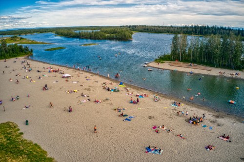 Aerial view of a sandy beach with people sunbathing and swimming in a river surrounded by trees and blue skies.