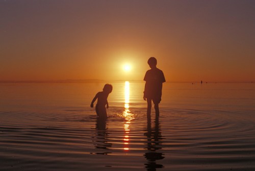 Two children play in shallow water at sunset, silhouetted against a vibrant orange sky.