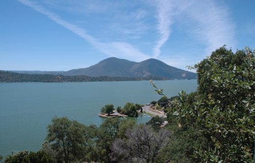 Scenic view of a lake surrounded by mountains and trees under a clear blue sky with wispy clouds.