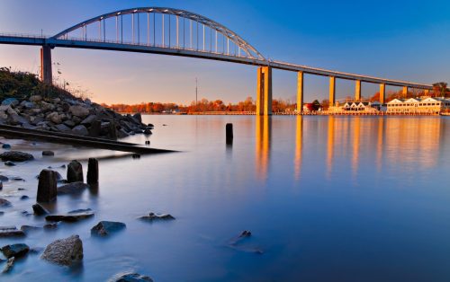 A serene river scene featuring a bridge, calm water, and autumn foliage reflecting in the sunset light.