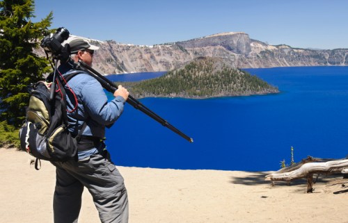 A photographer with a tripod stands by a scenic viewpoint overlooking a deep blue lake and forested island.