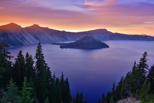 A serene view of Crater Lake at sunset, with a small island and surrounding mountains under a colorful sky.
