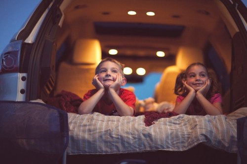 Two children resting in the back of a car, smiling and looking out, with soft lighting and cozy blankets around them.