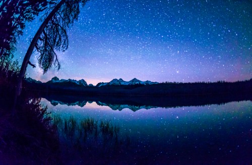 A serene night sky filled with stars reflects on a calm lake, surrounded by snow-capped mountains and trees.
