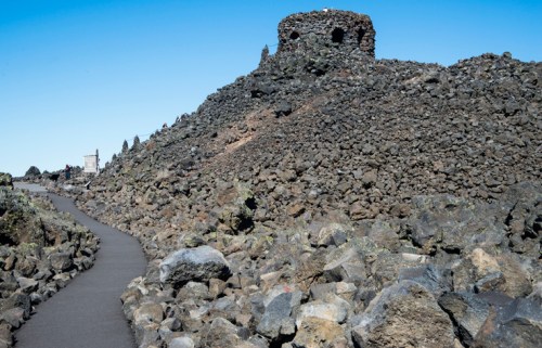 A rocky landscape with a winding path leading to a stone structure atop a hill under a clear blue sky.