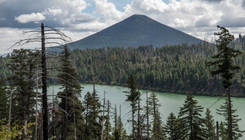 A scenic view of a mountain rising above a lake, surrounded by dense forest and cloudy skies.