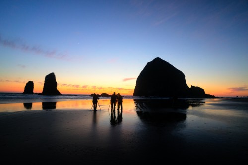 Silhouetted figures on a beach at sunset, with rocky formations and colorful sky reflected in the wet sand.