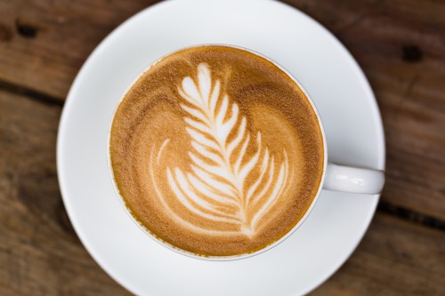 A close-up of a latte with intricate leaf latte art, served in a white cup on a wooden table.