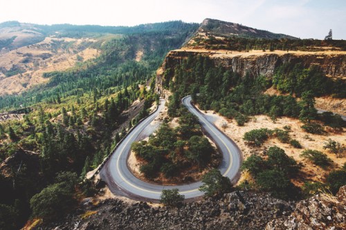 Winding road curves through a mountainous landscape with lush greenery and rocky cliffs under a hazy sky.