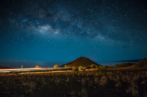 A starry night sky over a mountain, with a winding road illuminated by car lights in the foreground.
