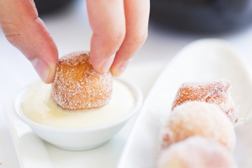 A hand dips a sugar-coated doughnut hole into a small bowl of creamy sauce.