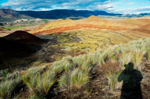 Vibrant, multicolored hills under a blue sky, with grassy foreground and a shadow in the lower corner.