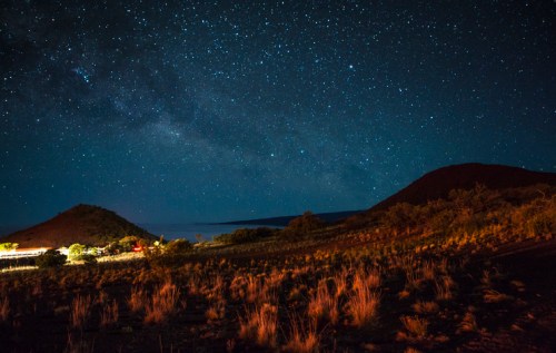 Starry night sky over a mountainous landscape, with a hint of light from a distant source illuminating the foreground.