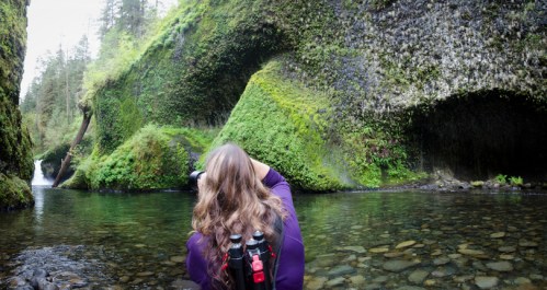 A person with long hair photographs a lush, moss-covered canyon near a tranquil stream and waterfall.