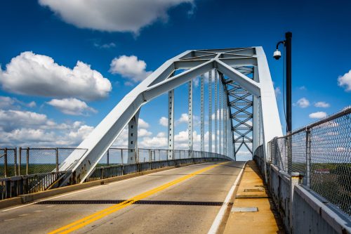 A white arch bridge spans a road under a blue sky with fluffy clouds, featuring a chain-link fence along the sides.