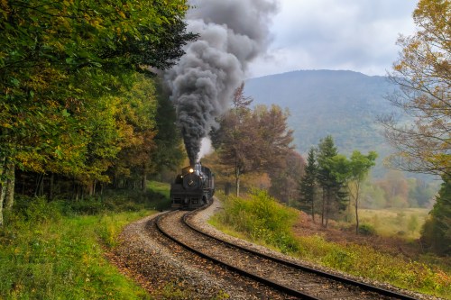 A vintage steam train chugs along a winding track through a colorful autumn forest, with smoke billowing into the air.
