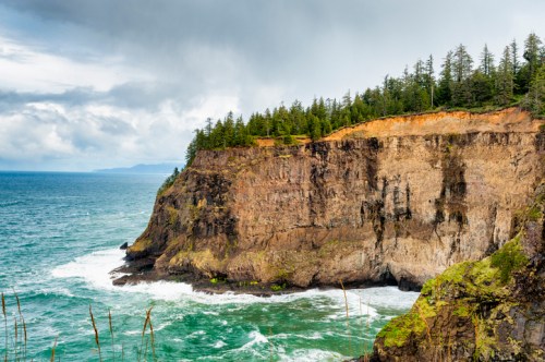 A rugged cliffside with trees overlooking a turbulent ocean under a cloudy sky.