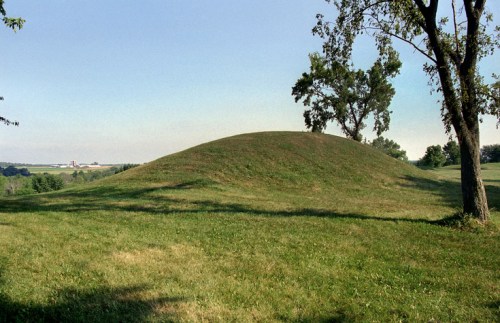 A grassy burial mound surrounded by trees, set against a clear blue sky and distant farmland.