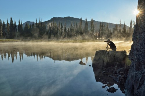 A photographer captures a misty lake scene at sunrise, surrounded by tall trees and mountains in the background.