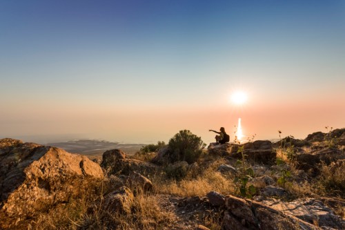 A person sits on a rocky hillside, pointing towards a sunset over a distant landscape.
