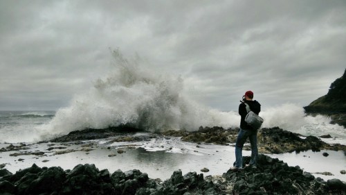 A person stands on rocky shore, capturing a large wave crashing against the rocks under a cloudy sky.