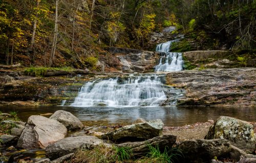 A serene waterfall cascades over rocky ledges, surrounded by autumn foliage and a calm pool below.