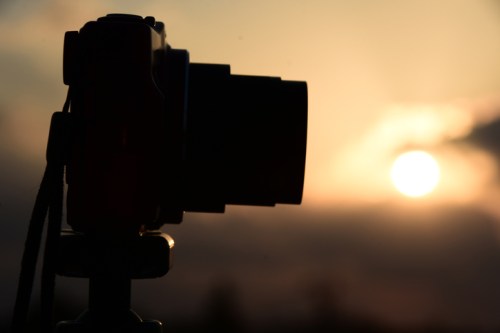 Silhouette of a camera on a tripod against a sunset backdrop with soft clouds.