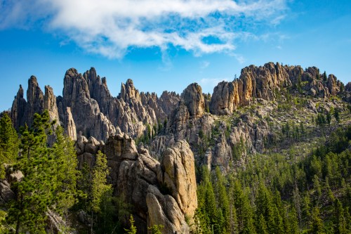 A rugged mountain landscape with towering rock formations and lush green trees under a blue sky with scattered clouds.