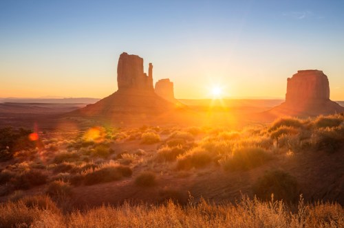Sunrise over Monument Valley, illuminating red rock formations and desert landscape with warm golden light.