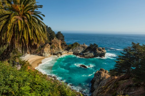 A scenic coastal view featuring turquoise waters, sandy beach, and lush palm trees against a clear blue sky.