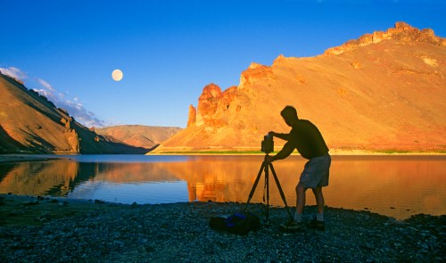 A silhouette of a photographer setting up a camera by a tranquil lake at sunset, with a full moon in the sky.