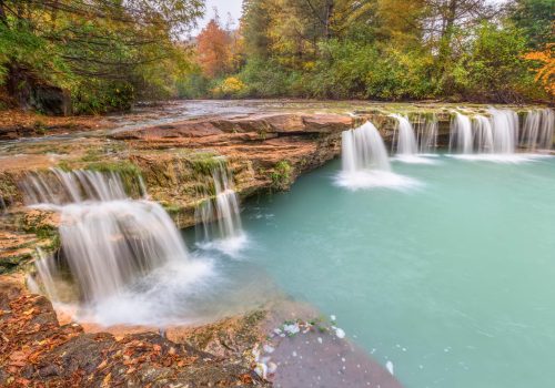 A serene waterfall cascades over rocky ledges into a turquoise pool, surrounded by autumn foliage.