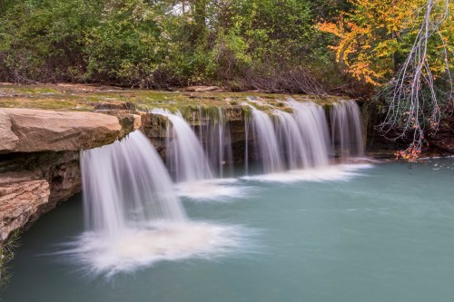 A serene waterfall cascades over rocky ledges into a calm, turquoise pool, surrounded by lush greenery and autumn foliage.